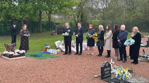 Seven smartly dressed men and women stand in a row watching a woman conduct a funeral service in a cemetery. The celebrant is standing above a hole in the red gravel floor which has been marked out with green felt. Several fo the people are holding flowers