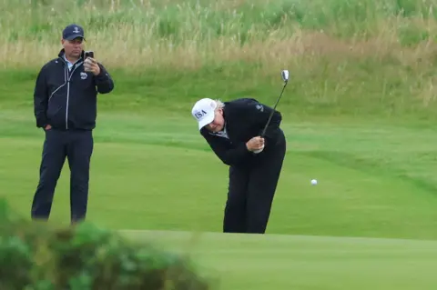 Reuters Donald Trump, wearing navy trousers and a navy jacket with a white "USA" cap, is doubled over as he strikes a golf ball.