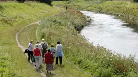 Picture of the Mersey River - walkers walk along the side of the river.
