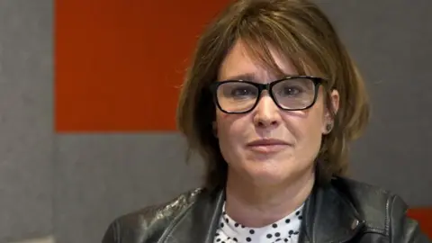 Carly Brookfield, who has short brown hair, glasses with black rims and is wearing a black leather jacket over the top of a white and black spotted T-shirt. She is smiling.