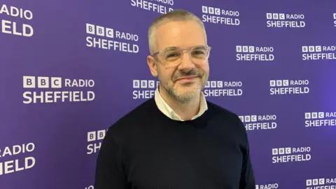 A man with close cut grey hair and stubble is wearing glasses, a white shirt and a blue jumper. He is standing in front of a purple wall with the words BBC Radio Sheffield
