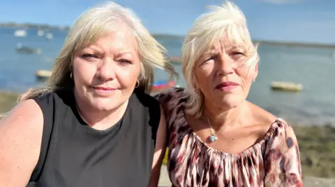 Nikki Fox/BBC A head and shoulders shot of Lisa Morris and Melanie Leahy standing outdoors on a sunny day looking at the camera. There is water and small boats behind them. Lisa has long hair and is wearing a black top. Melanie also has long hair and is wearing a leopard-skin top. 