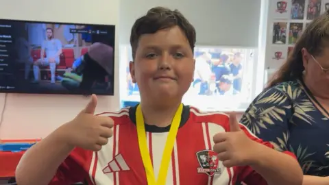 Oliver looking towards the camera with his thumbs up. He is smiling and is wearing a red and white striped shirt. There is a Exeter City FC badge on the right side of the shirt. Behind him is a TV and a woman stood to his right.