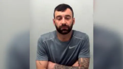 Essex Police Sidney Kemp has short black hair and a beard. For this picture, taken in police custody, he is wearing a grey T-shirt and looking downwards.