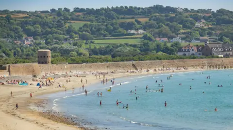 Visit Guernsey The beach and sea at Vazon Bay is full of beachgoers on a sunny day with green fields, trees and a few houses on the hills in the background.