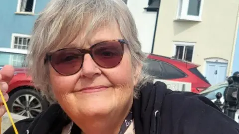 A head and shoulders shot of Sue Beacock wearing sunglasses. She has mid-length grey hair and is wearing a black jumper.
