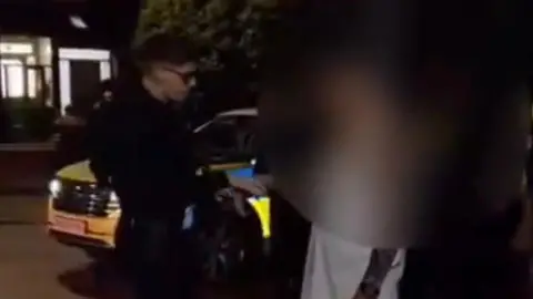 A male police officer with light blonde hair and glasses takes the right hand of a man, who is wearing a white t-shirt, and places it in handcuffs. A police patrol car is visible behind the officer. 