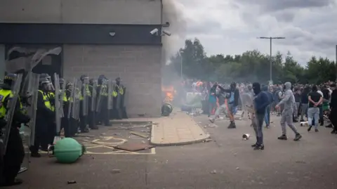 Getty Images A police line on the left, a crowd of people with hoods and face coverings on the right, a fire in the middle set next to a hotel building.