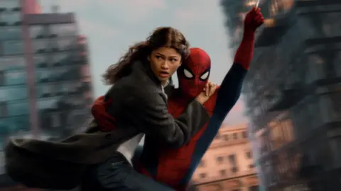 Spider-Man swings through the city on web whilst holding MJ, played by Zendaya.