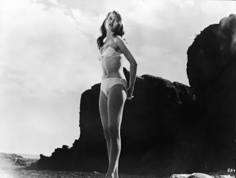 Atlantis Films/Pictorial Parade/Courtesy of Getty Images French-born actor Brigitte Bardot wears a white bikini and stands on a rocky beach in a still from the film, 'The Girl in the Bikini', directed by Willy Rozier, 1958.
