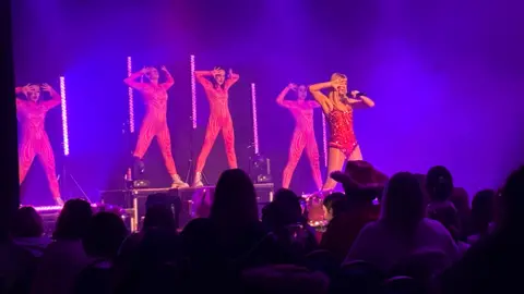 A woman who is dressed like Taylor Swift - in a red sequinned bodysuit - stands at the front of a stage with a microphone. Four backing dancers can be seen posing behind her. The backs of people's heads in the audience can be seen in the foreground. 