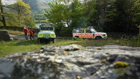 Two white Land Rovers with orange and green liveries parked next to a road. Two people in orange coats are starting to walk away form the vehicles, with a lake and trees behind them