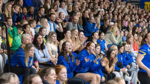 Josh Le Feuvre A large crowd sitting in stands – mainly women. Many are either looking happy and clapping, or uncertain, as the match progresses.