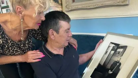 David Spereall/BBC Kristina and Alan Hunter observe a framed monochrome photo of Paul Hunter. Alan is sat down. Kristina stands behind him with her arms on his shoulders.