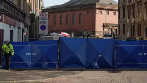 A large blue police cordon blocking the entrance to a street. Police officers are visible around it.