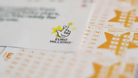 PA Media Close-up shot of EuroMillions logo - a drawing of fingers crossed with yellow stars behind it - on top of a sheet of yellow numbers.