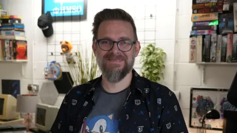 Peter Leigh standing inside a room filled with retro technology and gadgets. He is looking directly at the camera and smiling. He is wearing a grey Sonic the Hedgehog character T-shirt with a shirt over the top and a pair of glasses.