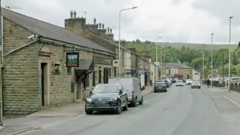Google Google streetview showing old stonebuilt two-storey houses or properties lining the street with cars parked in front. Pennine hills and trees are seen in the backdrop.