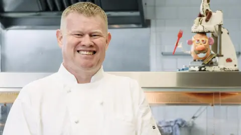 Andrew Crook is pictured standing in a professional kitchen. He is wearing chef whites and is smiling at the camera. He has short, blonde hair. On the counter behind him there is a Mr Potato Head toy. 