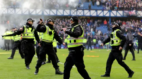Several police officers raise their batons as they stand on the pitch at Ibrox