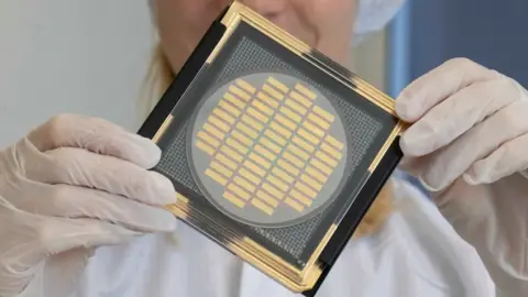 Getty Images A scientist in "clean room" gear holds up a large computer chip wafer the size of a fist