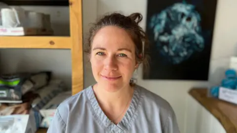 Ruth Geegan is smiling into the camera. She has brown hair, blue eyes and is wearing grey scrubs. She is standing in a vet surgery.