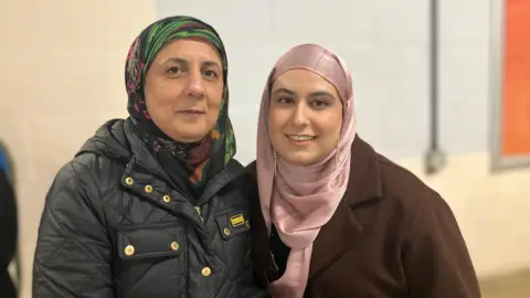 Shariqua Ahmed/BBC Lubna, a woman wearing a colourful scarf and a black coat and her daughter in a pink scarf and a brown coat, smiling for camera. 
