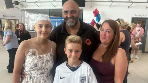 Philippa Goymer/BBC Keira Gawthorpe smiling into the camera alongside her family. She is wearing a white patterned headscarf and a matching white and purple floral dress. She has what looks like a cannula or another medical device on her chest which is just about visible. Her dad is to the right and is bald with a dark and grey beard. He is wearing a black polo shirt with an orange logo. In front of him is a young boy with blonde, short hair. He is wearing a white and blue England football shirt. Gawthorpe's mum is on the right has has long, brown hair and is wearing a purple vest top. She is looking to the right and looks like she has been crying.