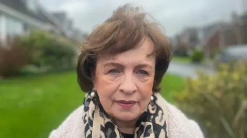 Diane Dodds has short, brown hair. She is looking at the camera with a neutral expression on her face. She is wearing an animal print scarf and a furry, cream coat. She is standing outside a housing estate, but the background is blurred.