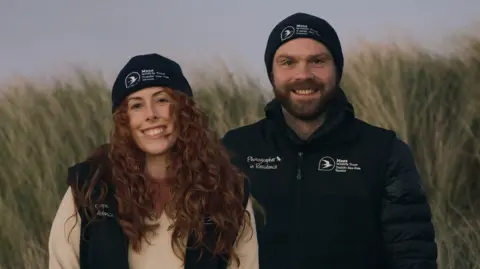 Adam Morgan/Ciara Kaneen Ciara (left) and Adam stands smiling for a photo. They're stood in long grass wearing warm clothing. Ciara has long, curly ginger hair whilst Adam has short hair and facial hair.