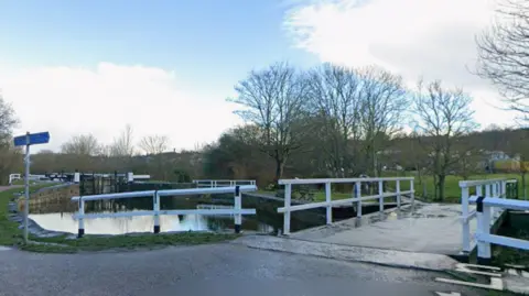Google A view of the canal at Saltaire. A road bridge crosses it with white railings and lock gates in the distance on the right.