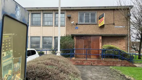 An outside view of the former South Ockendon police station, with a "for sale" sign on the side of the building. The front door has been boarded up and there is graffiti on it.