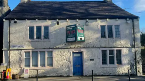 A large white building with a blue door and a dark slate roof. It has a large To Let sign fixed to the building.