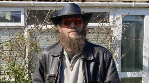 A man in sunglasses with a long brown beard, wearing a cowboy hat and a black leather jacket, stands in front of twigs and a light green wall with white windows.