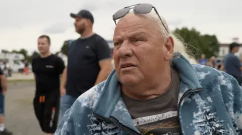 Gerd is an older gentleman with long white hair in a low pony-tail. He is wearing aviator sunglasses and a fleece decorated with wolves and arctic landscape.