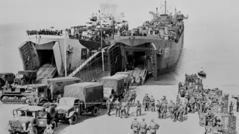 Getty Images Military vehicles being loaded aboard two tank landing ships at a beach. There are dozens of military personnel around the ships. The image is black and white