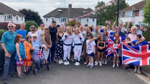 Damian Derrick/BBC Olympic medallists Kate Shortman and Isabelle Thorpe stand in their Team GB tracksuits among a group of family and friends on their return to Bristol. People in the group are holding up Union flags and smiling at the camera, with houses in the background