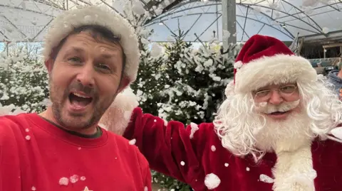 Combe Garden Centre manager James Trevett cheers as he stands next to Father Christmas in front of Christmas trees at the shop. Fake snow made from bubbles is coming down from the sky. Mr Trevett is wearing a red jumper and a Christmas hat.