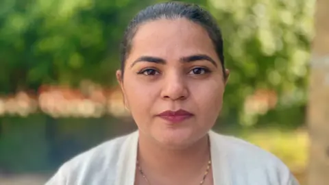 Stephen Huntley/BBC A woman with dark hair and dark eyebrows looks directly into the camera lens. She wears a white cardigan and necklace. Her lips are slightly upturned into a smile. She is standing outside. The trees and greenery behind her have been blurred.