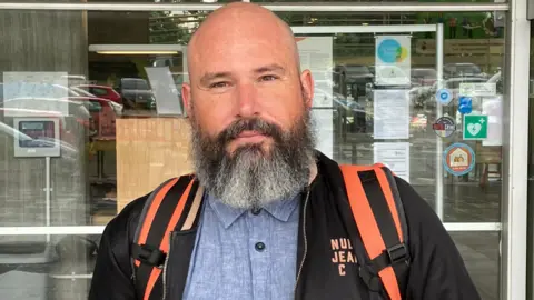 John Brown, a bald man with a medium-length grey and white beard, wearing a black jacket and with black and orange rucksack straps over this shoulders in front of a window 