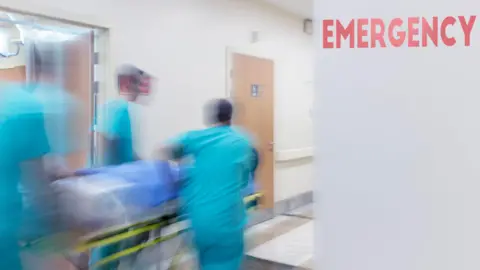A blurred action shot of three medical professionals wheeling a patient on a trolley down a hospital corridor. The medics ae wearing light blue scrubs. There's a red sign reading 'emergency' on a grey wall. 