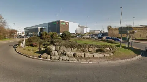 The stones on the roundabout. The stones are displayed in three tiers and there is shrubbery growing on top of the highest set. Grass covers the rest of the roundabout. An Asda supermarket is in the background.