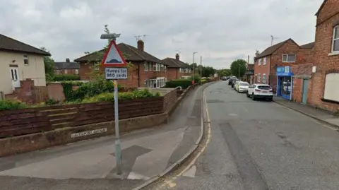 A street, with houses either side and a sign warning motorists about speed bumps. The house on the left hand side of the image has a wooden slatted fence around it, and there are a number of parked cars on the right hand side of the image.