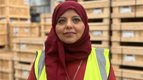 Shaista Aziz is wearing a red hijab as well as a yellow hi-vis jacket over a ted top. She is standing in front of large wooden pallets.