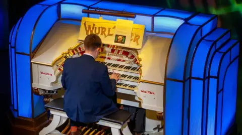 Damon Cox/Wurlitzer Heritage Trust Oscar who is playing the organ wearing a suit.