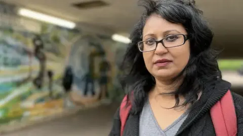 EMMA BAUGH/BBC Karima Shah is looking at the camera. She is wearing a grey T-shirt and a black jacket, looking into the camera. She is photographed standing in front of a colourful mural painted on a wall of an underpass. 