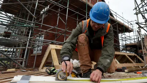 A man on a building site, with red-brick houses surrounded by scaffolding in the background. The man has a beard and wears glasses and a blue helmet. He is wearing a high-vis jacket and protective clothing. He is crouched on the ground with a metal tape measure extended on a strip of material in front of him.