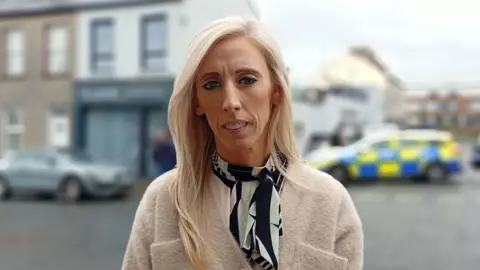 Carla Lockhart, a woman with long, straight, blonde hair, looks directly at the camera with a serious expression while standing close to the scene of the car fire in Portadown. She is wearing a beige cardigan with large front pockets and black and pale green scarf tied around her neck. The background is out of focus but there are houses and a parked police car behind her.