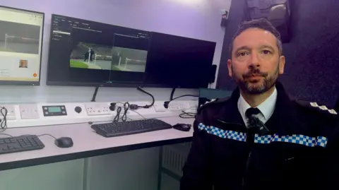 A male police officer sits in front of computer screens which shows live camera feed of a road and people walking