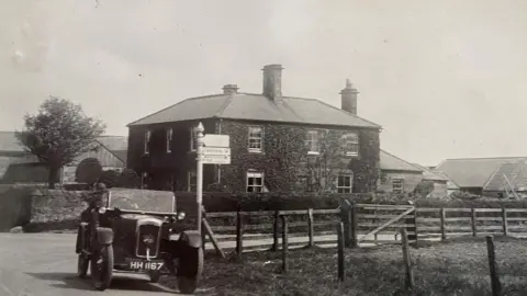 Martin Beal A black and white image of Charlton Mires taken in the 1920s with a car of the era being driven outside. The house is a large Georgian one with three chimneys and there are farm buildings behind and a wooden fence in the foreground. It sits on a junction with an old road sign.